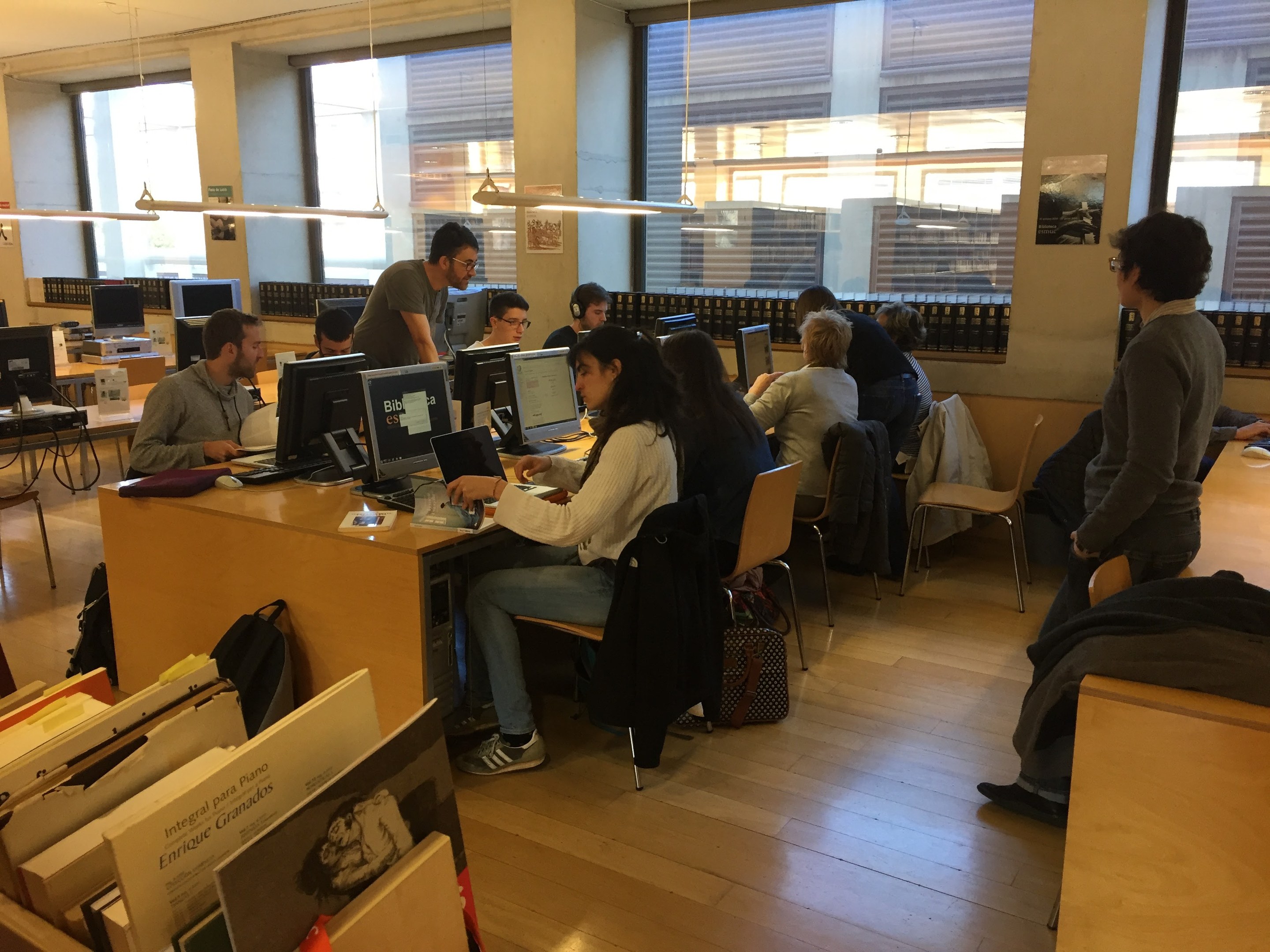 Gruppe von Menschen, die an Computern auf Schreibtischen in einer Bibliothek mit Bücherregalen, Monitoren, Tastaturen, Mäusen, Taschen auf dem Boden, Deckenlampen und Glasfenstern im Hintergrund arbeiten.