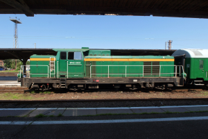 Ein grüner Diesel-Elektrolokomotive auf Eisenbahnschienen unter einer Brücke, umgeben von Steinen, Gras, einem Weg, Bäumen, Strommasten mit Drähten, einem Turm, Fahrzeugen und einem bewölkten Himmel.