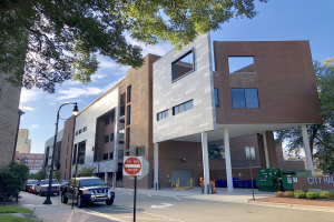 Großes mehrstöckiges Stadtzentrumsgebäude mit vielen Fenstern, umgeben von Straßeninfrastruktur einschließlich Pfosten, Lampen, Fahrzeugen, Schildern, Verkehrskegeln, Mülltonnen und Grünflächen unter einem bewölkten Himmel.