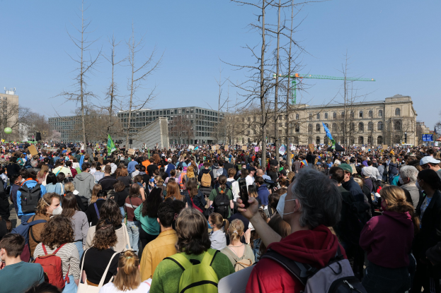 Große Menschenmenge bei einer Klimaprotestkundgebung vor einem Gebäude mit Bäumen und einem klaren Himmel im Hintergrund, viele halten Schilder und tragen Taschen.