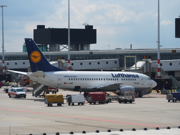 Lufthansa Airbus A320-200-Flugzeug auf dem Frankfurter Flughafen geparkt mit Bodenfahrzeugen und Wagen im Vordergrund, einem Gebäude und Laternen im Hintergrund unter einem klaren blauen Himmel.