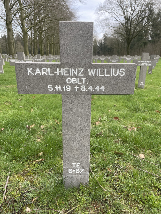 Grab von Karl-Heinz Willis auf einem deutschen Friedhof, umgeben von Gras und welken Bl├Ąttern, mit Trees und einem klaren blauen Himmel im Hintergrund.