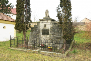 Ein steinerner Gedenkstein auf einer grünen Wiese, umgeben von einem Metallzaun und geschmückt mit Blumensträußen, mit Bäumen, Häusern und einer Wand im Hintergrund unter einem klaren blauen Himmel dient als Holocaust-Gedenkstätte in Sibiu, Rumänien.