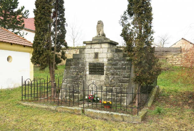 Ein steinerner Gedenkstein auf einer grünen Wiese, umgeben von einem Metallzaun und geschmückt mit Blumensträußen, mit Bäumen, Häusern und einer Wand im Hintergrund unter einem klaren blauen Himmel dient als Holocaust-Gedenkstätte in Sibiu, Rumänien.