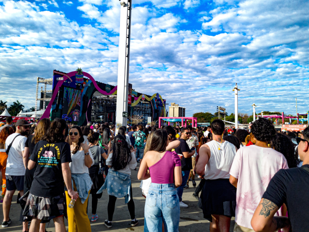 Eine Menschenmenge, einige mit Mützen und Schutzbrillen, steht vor einem Karneval mit Pfählen, Bäumen, Gebäuden und einem bewölkten Himmel im Hintergrund.