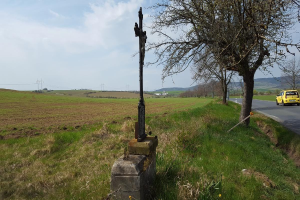 Ein gelbes Auto fährt auf einer Straße neben einem Baum und einem Straßenkreuz, mit Gras, Blumen, Hügeln und einem bewölkten Himmel im Hintergrund.