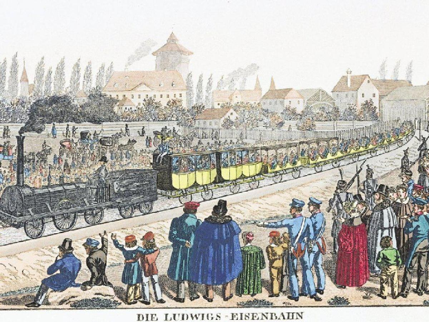 Eine Gruppe von Menschen steht um einen Zug auf einem Gleis, einige sitzen im Zug und andere stehen davor, vor Gebäuden und Bäumen, mit Text 'Ludwigs-Eisenbahn, die erste Bahnstation in Deutschland' unten.