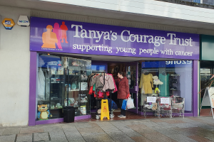 Ein Schaufenster mit einer Tafel, auf der "Tanyas Mutvertrauen" steht, mit einer Person davor, Kleidung, Teddybären, Puppen, einem Müllcontainer, einer Dose, einer Pflanze und einer Tafel mit Text auf der rechten Seite.