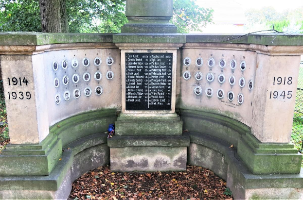 Ein Holocaust-Gedenkmonument in einem Berliner Jüdischen Friedhof, das eine Text- und Nummernwand an seiner Wand hat, umgeben von Bäumen, einem Zaun und verstreuten trockenen Blättern.