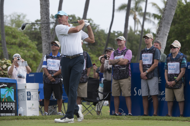 Ein Mann in einem weißen Hemd, schwarzen Hosen und einer blauen Mütze schwingt einen Golfschläger auf einem grünen Feld, mit Zuschauern und Kameras im Hintergrund und Bäumen und einem klaren Himmel.