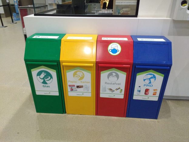 Vier bunte Recycling-Eimer in einer Reihe auf dem Boden, mit einer Arbeitsplatte, einem Monitor und anderen Gegenständen im Hintergrund.