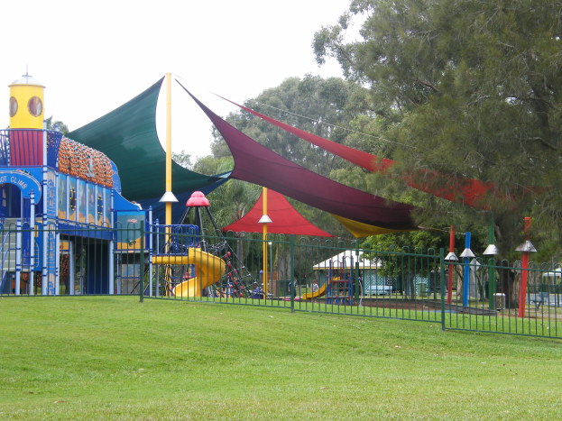 Ein Park mit einem Spielplatz, der von einem Metallzaun umgeben ist, mit Kindern, die auf Geräten wie einer Rutsche und Stangen spielen, umgeben von grünem Gras, Bäumen und einem klaren blauen Himmel.