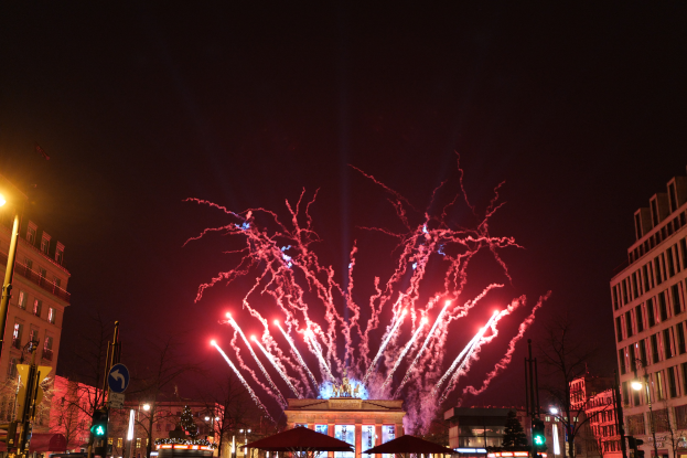 Eine belebte Stadtstraße in Berlin an Silvester, voller Menschen, Fahrzeuge und Gebäude, erleuchtet von Feuerwerk und Gebäudelichtern, die eine festliche Stimmung schaffen.