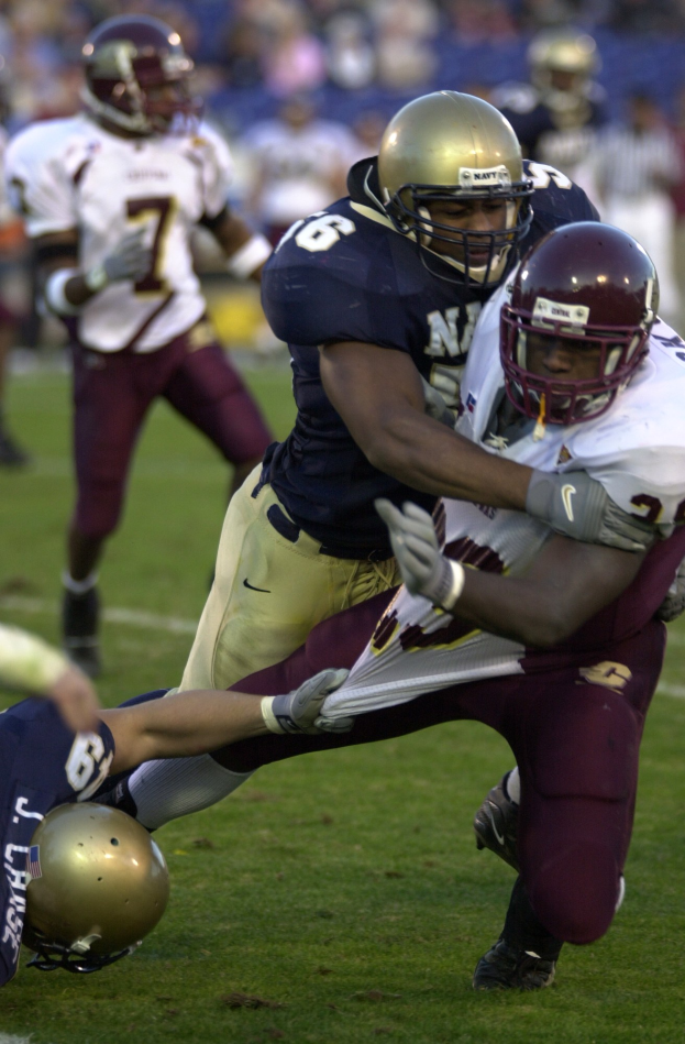 Zwei Fussballspieler in Helmen und Uniformen stoßen auf dem Feld zusammen, während andere zuschauen.