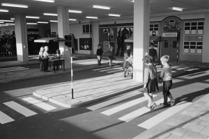 Ein Schwarz-Weiß-Foto von Kindern, die eine Straße an einer Ampel überqueren, mit Menschen, die stehen und sitzen, einer Verkehrsampel im Vordergrund und Säulen und Lampen im Hintergrund.