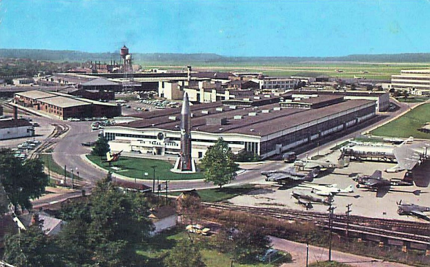 Alte Schwarz-Weiß-Luftaufnahme eines Flughafens mit geparkten Propellerflugzeugen, Terminalgebäuden, umliegenden Straßen, Fahrzeugen, Strommasten, Grasflächen, Bahnschienen, fernen Hügeln und einem klaren Himmel, mit gedrucktem Text entlang der unteren Kante.