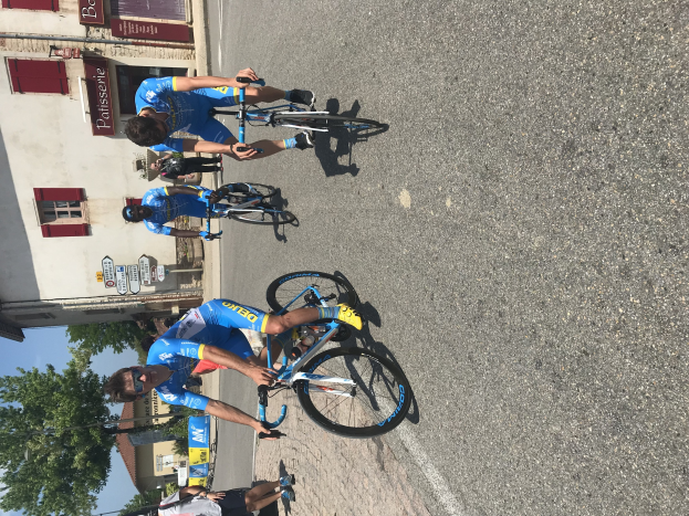 Eine Gruppe von Radfahrern fährt eine Straße entlang an einem Gebäude mit Fenstern und Schildern vorbei, mit Bäumen und einem klaren Himmel im Hintergrund und Zuschauern, die auf der Straße darunter stehen.