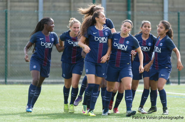 Gruppe junger Frauen beim Fußballspielen auf einem Rasenplatz mit Maschendrahtzaun und einer Wand im Hintergrund, Text unten rechts lautet "Paris Saint-Germain Frauenfußball".