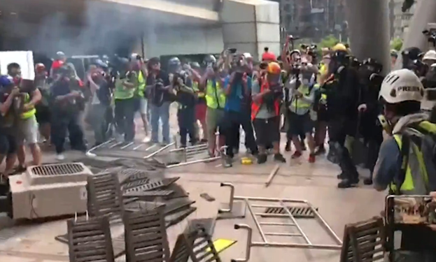 Menschen vor einem rauchenden Gebäude mit Helmen und Jacken, andere mit Kameras, mit verstreuten Metall-Objekten und Stadtgebäuden im Hintergrund.