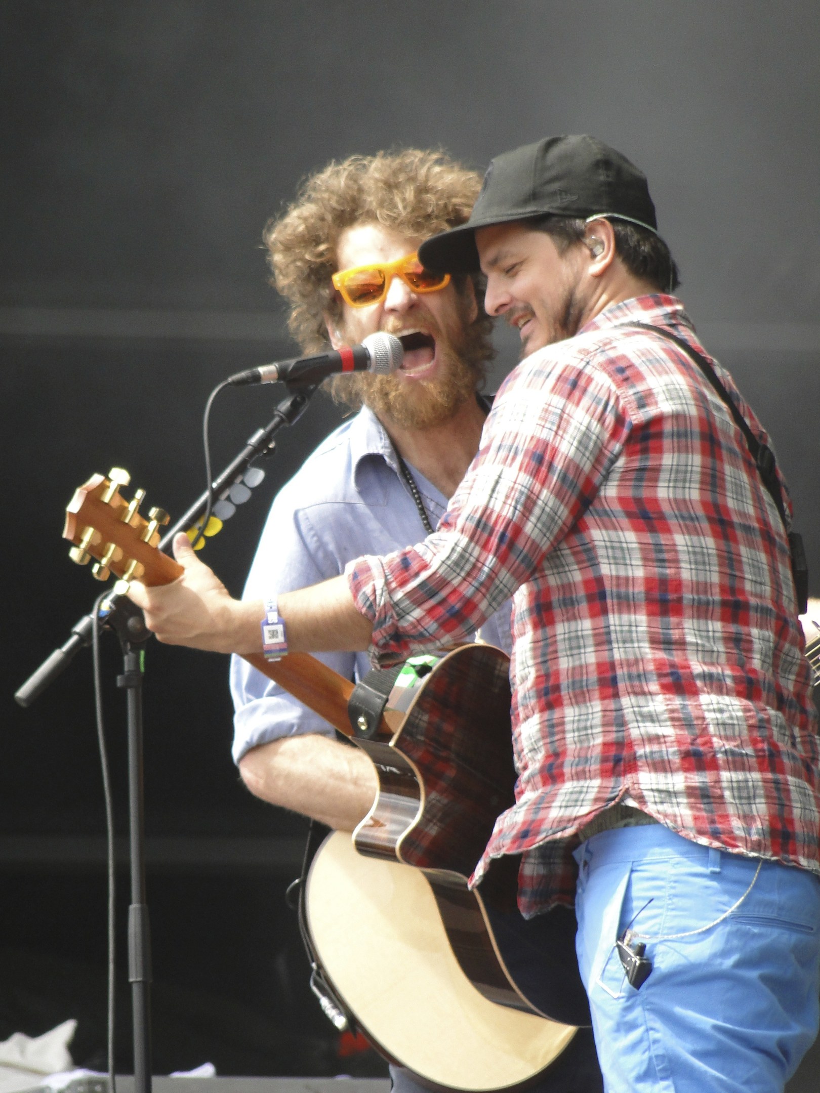 Zwei Männer auf der Bühne, einer in einem rot-blau karierten Hemd mit blauen Hosen, der andere mit einer schwarzen Mütze und einer Sonnenbrille, beide singen in Mikrofone und spielen Gitarren.