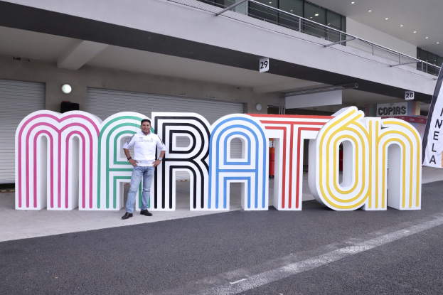 Ein Mann steht am Start eines Marathons vor einem großen "Marathon"-Schild, mit einer Fahne rechts und einem Gebäude mit Lichtern, Geländern und Fensterläden im Hintergrund.