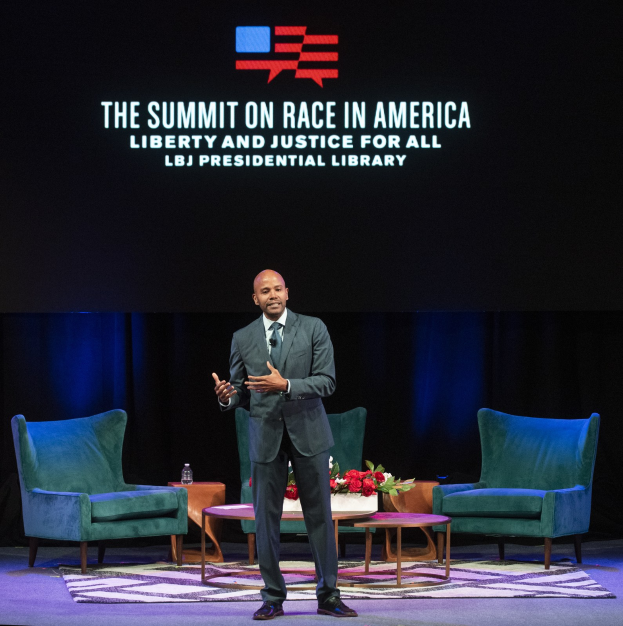 Ein Mann in Blazer, Krawatte und Hemd spricht auf der Bühne bei der Veranstaltung "The Summit on Race in America: Liberty and Justice for All" in der LBJ Presidential Library, mit zwei Stühlen, einem Tisch mit Blumenstrauß und Flasche, einem Vorhang und einem Bildschirm dahinter.