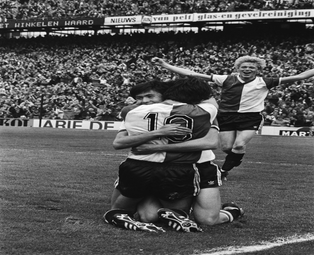 Drei Fußballspieler feiern ein Tor auf dem Boden, mit jubelnder Menge und schwenkenden Fahnen in einem schwarz-weißen Stadion-Setting.