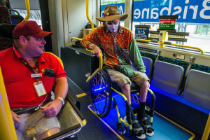 Zwei Männer im Rollstuhl in einem Bus, einer trägt ein rotes T-Shirt und eine Kappe, der andere ein orangefarbenes Hemd und einen braunen Hut, beide tragen Masken und ID-Karten, mit gelben Stangen und Glasfenstern, durch die Bäume, Gebäude und der Himmel zu sehen sind.