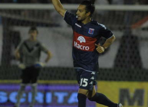 Ein Fußballspieler feiert auf dem Boden nach einem Tor, mit einer jubelnden Menge und einer Person im Hintergrund und einer Tafel mit Text dahinter, alles mit einem unscharfen, dynamischen Hintergrund.