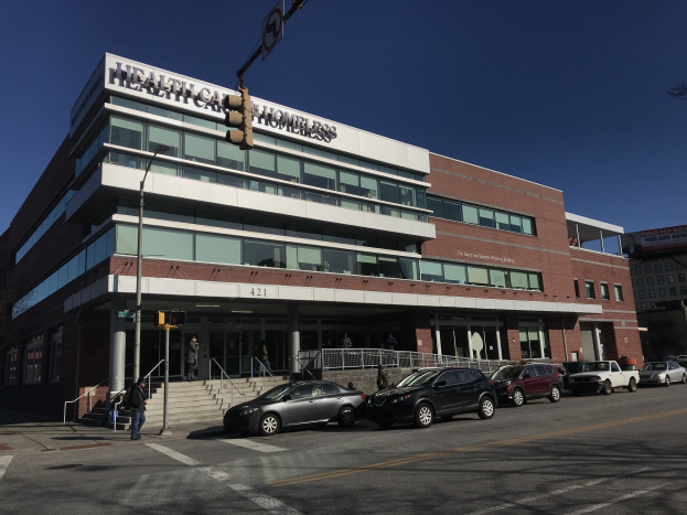 Großes Gebäude als New York City Health Care Homeless Center beschriftet mit parkenden Autos, Fußgängern, Straßenmöbeln und Bäumen unter einem klaren Himmel.