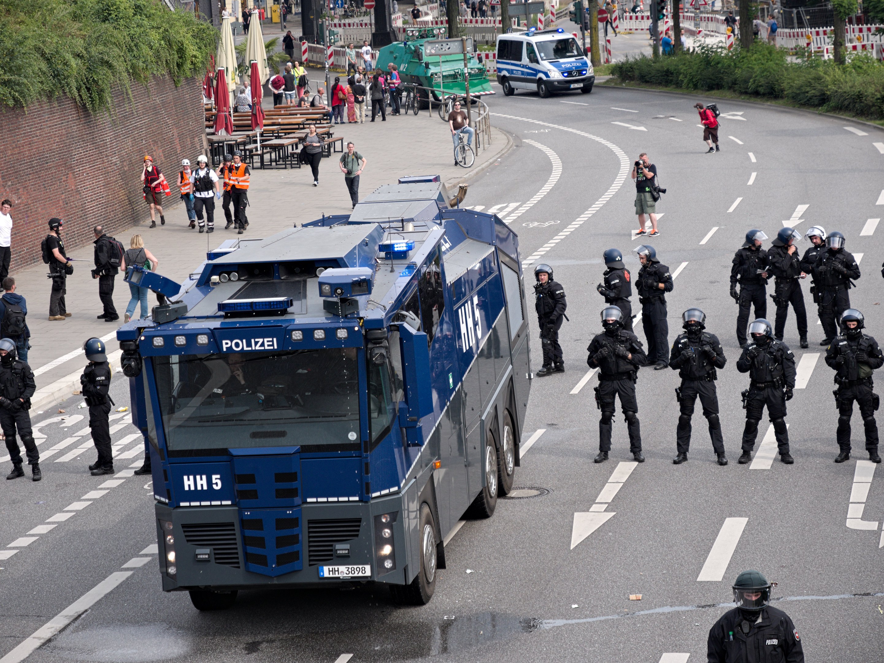 Gruppe von Polizisten vor einem gepanzerten Fahrzeug auf einer Stra├če mit bewaffneten Individuen und urbaner Umgebung, einschließlich Geb├Ąuden, Verkehrszeichen und Vegetation.