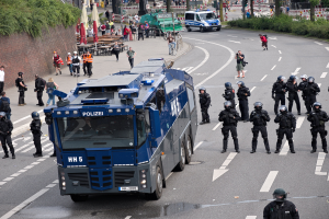 Gruppe von Polizisten vor einem gepanzerten Fahrzeug auf einer Stra├če mit bewaffneten Individuen und urbaner Umgebung, einschließlich Geb├Ąuden, Verkehrszeichen und Vegetation.
