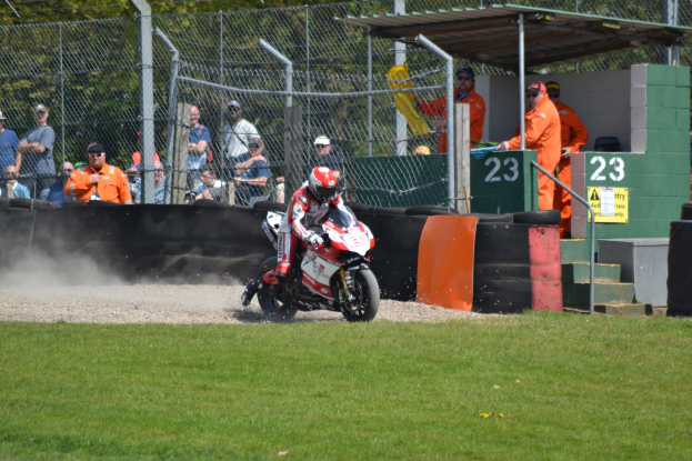 Ein Motorradfahrer in Helm und Handschuhen fährt über ein grasbewachsenes Feld, mit Zuschauern in Mützen und Schutzbrillen, einem Zaun mit Bäumen, Treppen mit Geländern und einem nummerierten Schuppen im Hintergrund.