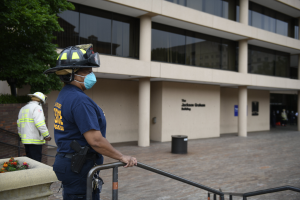 Ein Polizist in einer Gesichtsmaske und einem Helm steht vor einem Gebäude mit Glasfenstern, Säulen und Text an der Wand, mit einem Geländer im Vordergrund, einem Baum links und ein paar Menschen und einem Müllcontainer im Hintergrund.