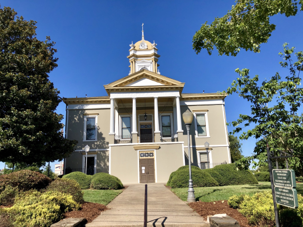 Großes Backsteingebäude mit einem Glockenturm, umgeben von Fenstern, Türen, Säulen, Geländern, einem Schild, Straßenlaternen, Bäumen, Gras und einem bewölkten Himmel, beschriftet als das wiedereröffnete alte Rathaus.