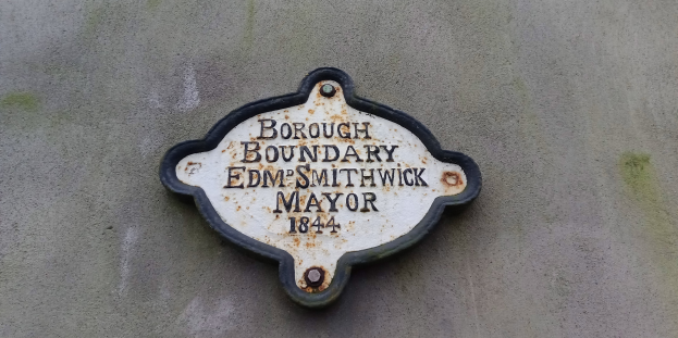 Metallschild an der Außenwand eines Gebäudes, das die Borough-Grenze markiert, mit "Edm Smithwick Mayor" in fetter weißer Schrift auf schwarzem Hintergrund.