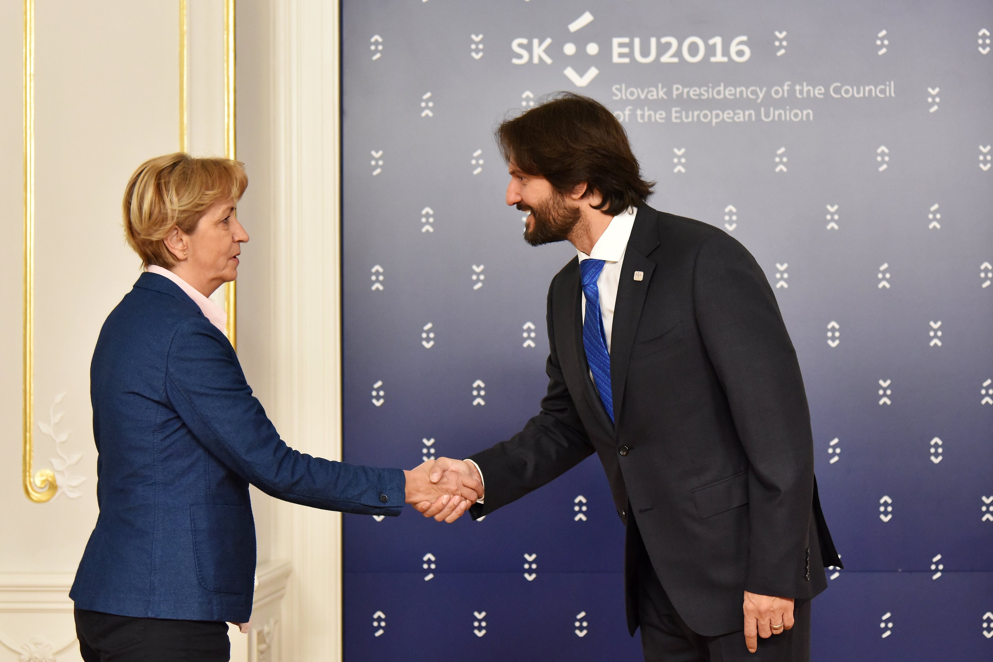 Bundeskanzlerin Angela Merkel und Bundeskanzler Joachim Gauck schütteln sich freundschaftlich die Hand vor einer Wand mit einem "EU-Gipfel"-Schild und einer Tür.