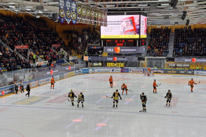 Eine Gruppe von Menschen mit Helmen und Hockeystöcken spielt ein Spiel auf einem Eisstadion mit einem Netz in der Mitte, umgeben von Texttafeln und einem großen Bildschirm im Hintergrund, mit einer Person mit Hut.