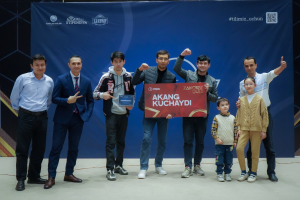 Gruppe von Menschen auf einer Bühne, die eine Tafel mit dem Text "Akang Kuchaydi - Zakovat Quiz" halten, mit einem Banner, Lichtern und anderen Gegenst├Ąnden im Hintergrund.