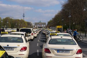 Eine lange Reihe von Taxis, die auf der Seite einer belebten Straße in Berlin, Deutschland, geparkt sind, mit Fahrzeugen, Radfahrern und Fußgängern, flankiert von Laternenpfählen, Bäumen und Gebäuden, einschließlich eines Bogens und eines Turms, unter einem bewölkten Himmel.