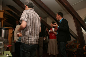 Mehrere Personen mit Tassen in der Hand, ein Becher und ein Tisch links, ein Dach oben und Stühle rechts.
