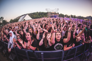 Eine große Menschenmenge auf einem Musikfestival mit erhobenen Händen vor einem Geländer, Bäumen, Gebäuden und einem klaren blauen Himmel im Hintergrund.