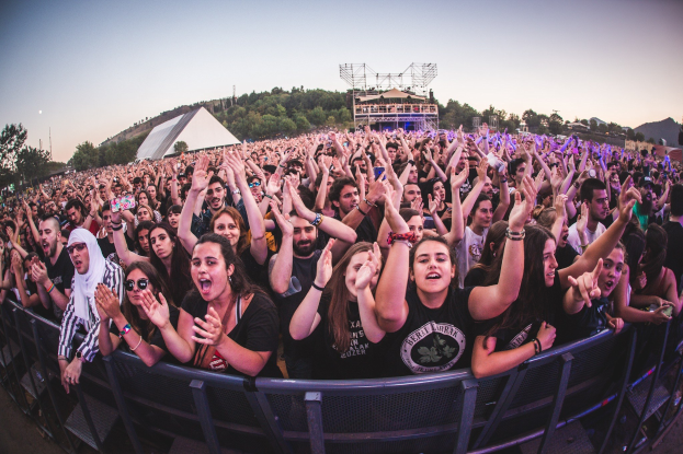 Eine große Menschenmenge auf einem Musikfestival mit erhobenen Händen vor einem Geländer, Bäumen, Gebäuden und einem klaren blauen Himmel im Hintergrund.