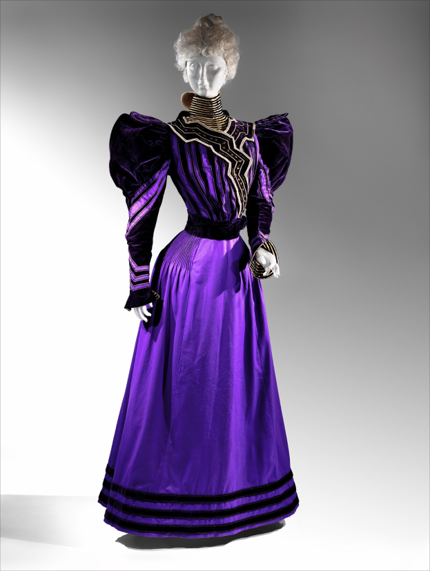 Eine Schaufensterpuppe trägt ein bodenlanges lila Abendkleid mit einer eng anliegenden Taille, einem weiten Rock, einem schwarzen und goldenen gemusterten Stoff, goldener Stickerei, Perlen und einer Spitzenborte an der Taille, kombiniert mit einem eleganten Hochsteckdutt und zarten Ohrringen.
