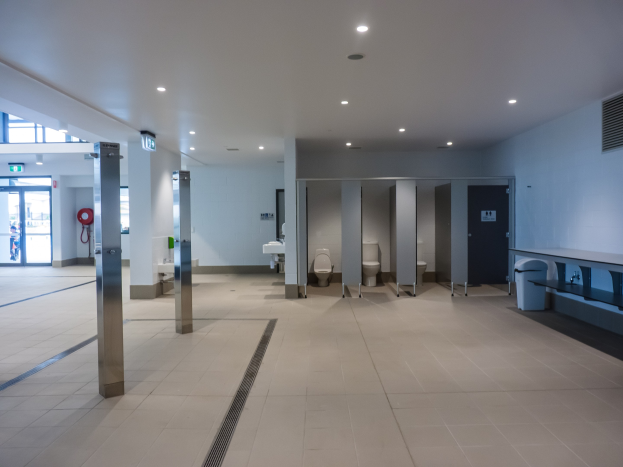 Große öffentliche Toilette mit mehreren Toiletten, Säulen, einem Mülleimer, einer Tür, einem Schild, einem Feuerlöscher und Deckenleuchten.