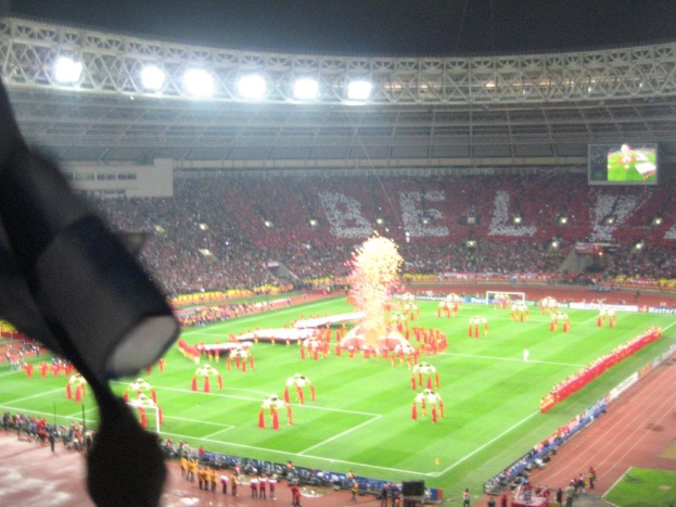 Ein Stadion mit Flutlicht, Eisen Gittern, Zuschauern, Personen am Boden, Werbetafeln, Anzeigetafeln, einer Überwachungskamera, Fahnen und Dekorationen.