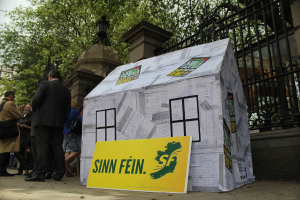 Ein Zelt und ein Werbeschild sind auf dem Boden platziert, während eine Menschenmenge daneben steht. Im Hintergrund sind ein Eisengitter, Säulen, Straßenlaternen, Bäume und der Himmel zu sehen.