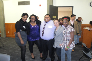 Menschen in der Mitte, ein Podium und Stühle rechts, eine Wand und eine Tür im Hintergrund.