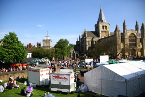 Historische Gebäude mit Bäumen davor, zusätzliche Gebäude und ein Turm im Hintergrund, ein öffentlicher Weg mit Geschäften und Menschen, die auf dem Gras in der Nähe sitzen.
