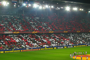 Ein Fußballspiel wird in einem großen, hell erleuchteten Stadion gespielt, mit Zuschauern auf dem Feld stehend und auf den Tribünen sitzend.